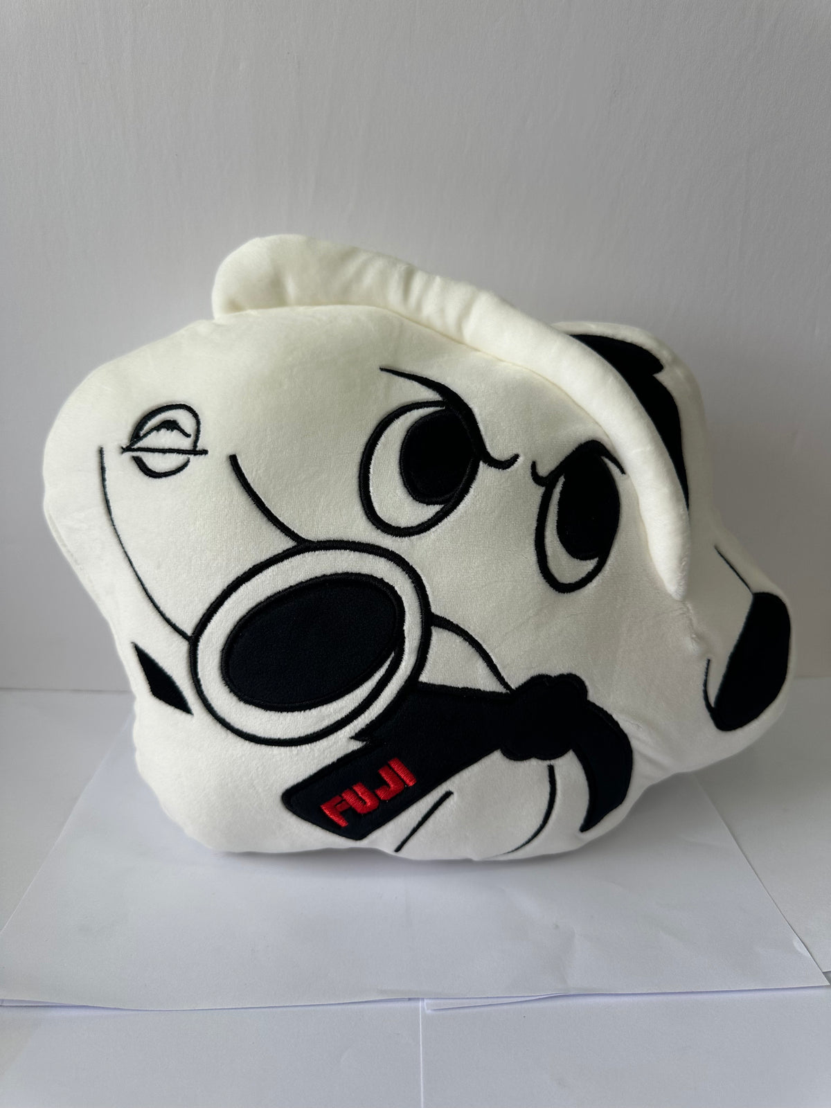 The Pochi Piro Pillow White features a soft white plush surface with bold black outlines, a playful cartoon face, large expressive eyes, and a black bar marked FUJI for a fun, eye-catching look.