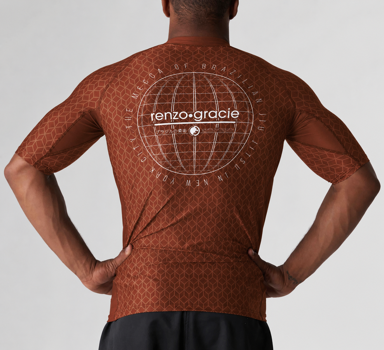 A person stands with hands on hips, wearing the Renzo Gracie Mecca Ranked Short Sleeve Rashguard. The brown patterned athletic shirt features a white globe graphic and "renzo gracie" text in circular lettering on the back.
