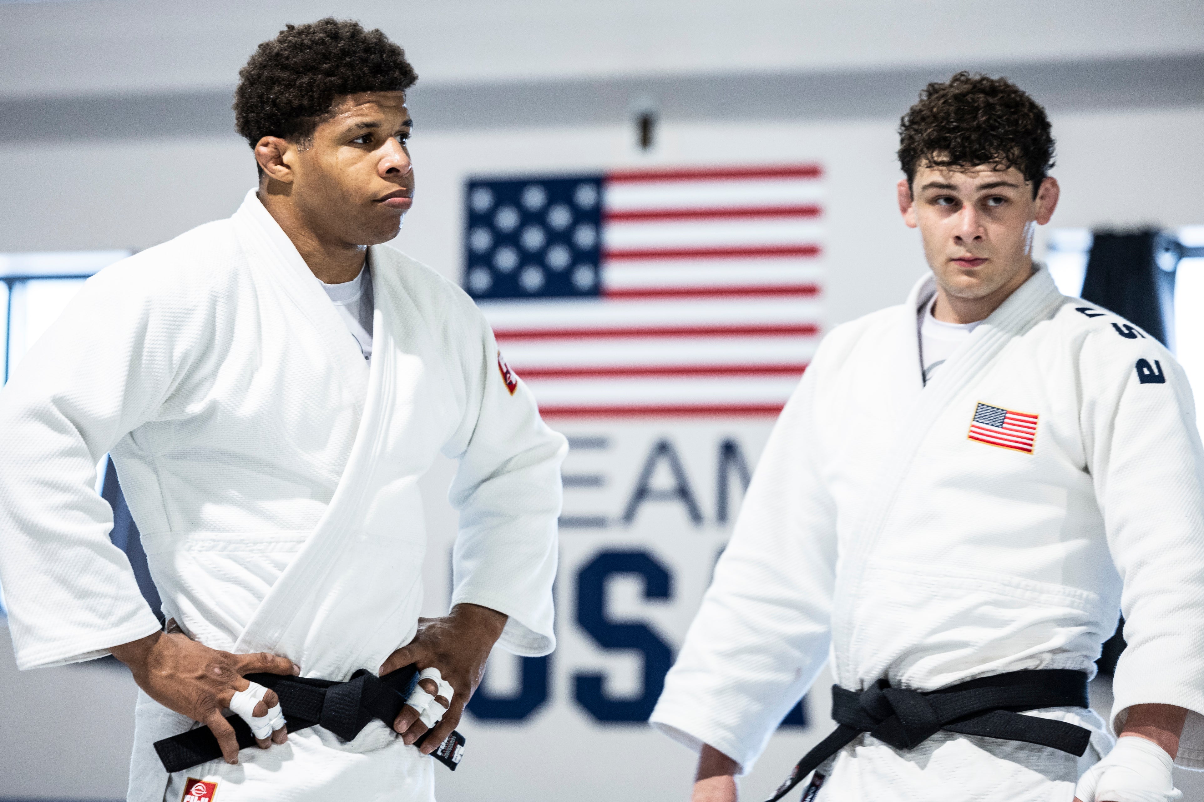 Two men wearing white jiu-jitsu uniforms stand side by side in a gym, with an American flag and the words “TEAM USA” visible in the background. Both look focused and attentive.