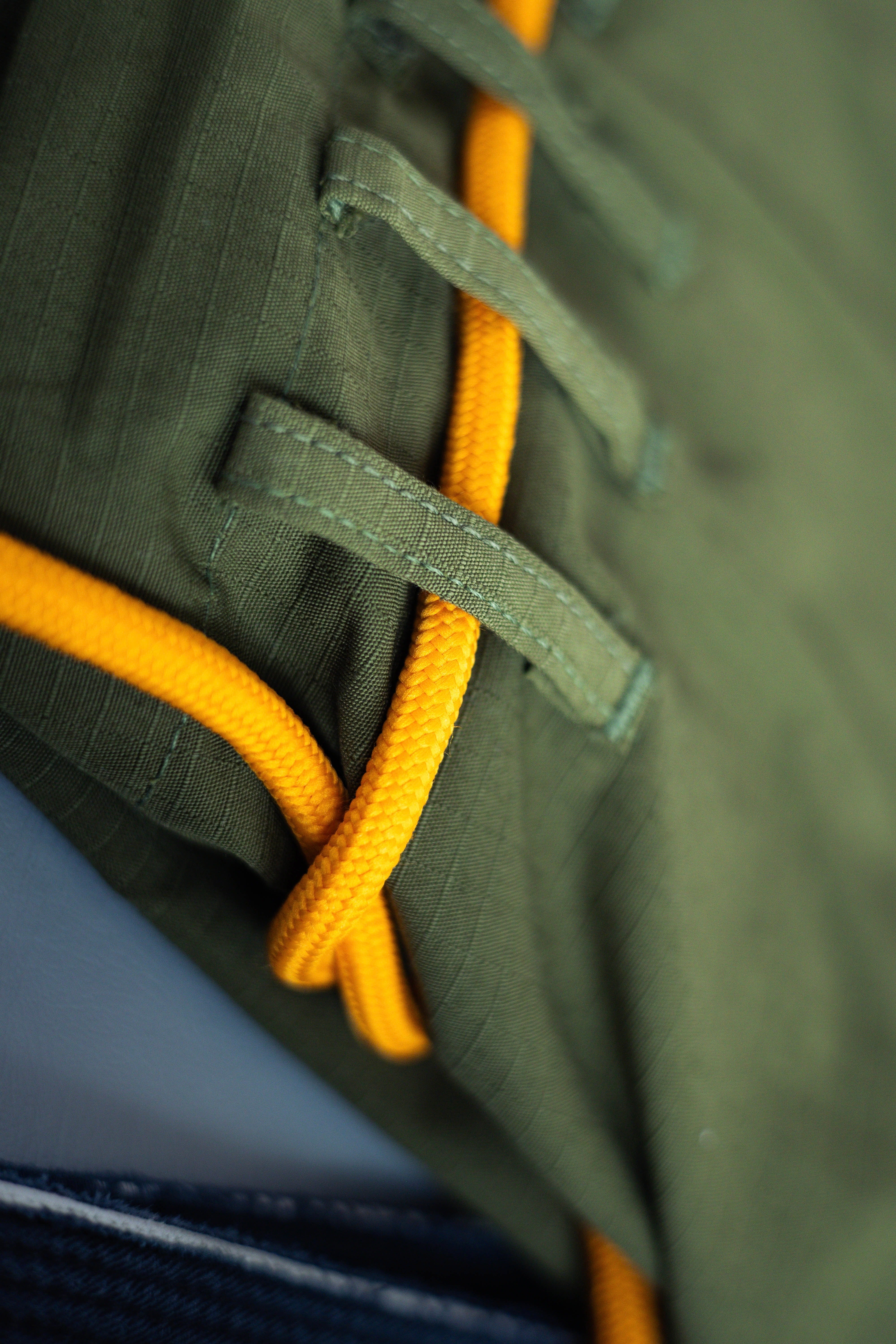 Close-up of the We Defy Ripstop BJJ Gi in Military Green, showing durable looped fabric with a bright yellow cord laced through, plus blue denim with white stitching at the bottom.
