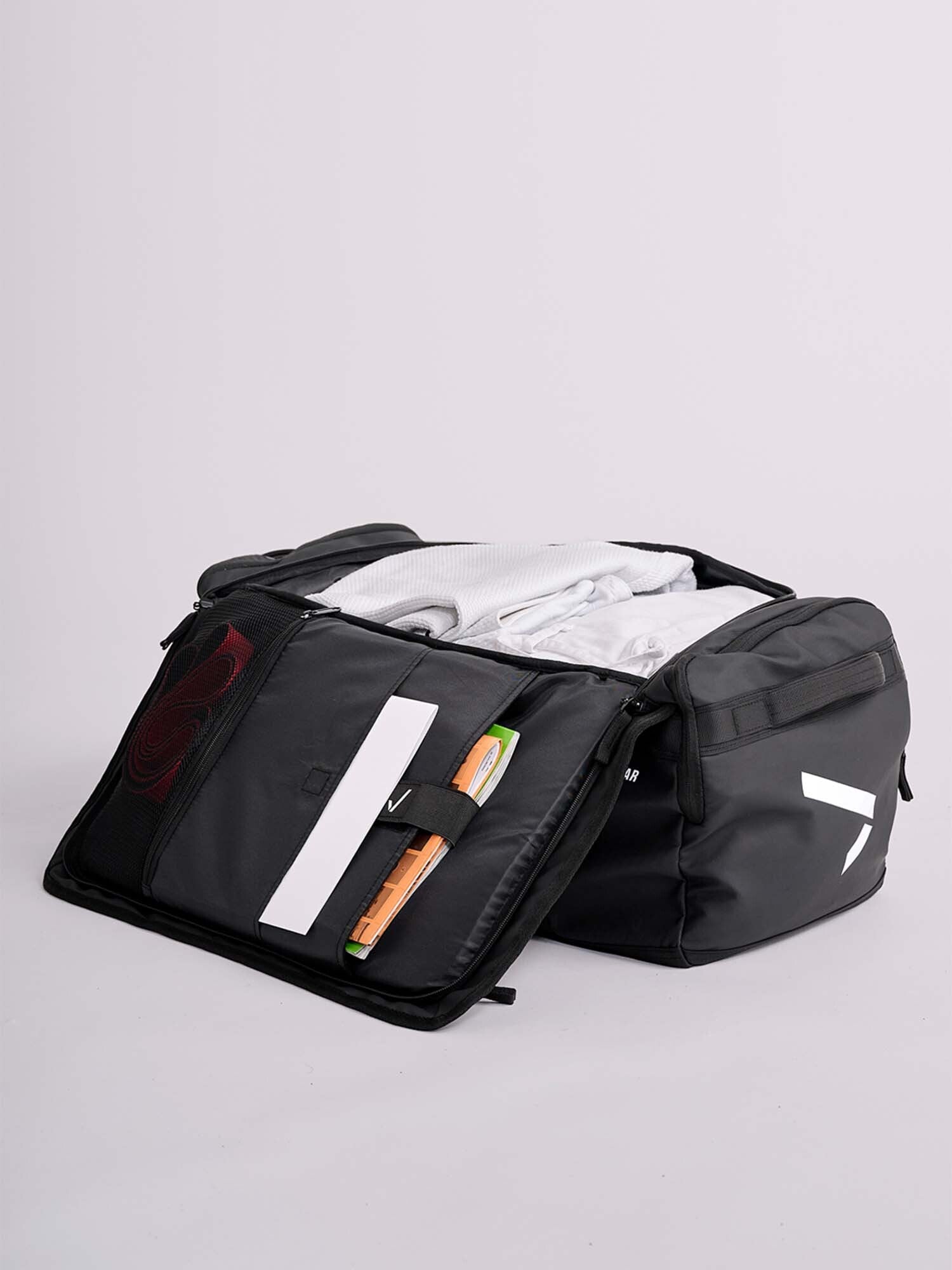 The IPPONGEAR 2 in 1 Sports Bag Black, unzipped and open on a light surface, shows neatly packed clothes and organized pockets with a notebook, pens, and small items.