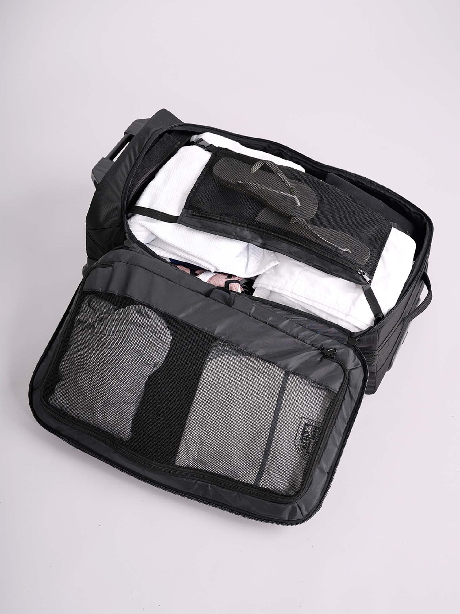 An open IPPONGEAR Travel Trolley Black L shows organized shirts, black flip-flops, and clothes inside. The lid features mesh compartments with neatly folded garments. The suitcase rests on a plain, light surface.