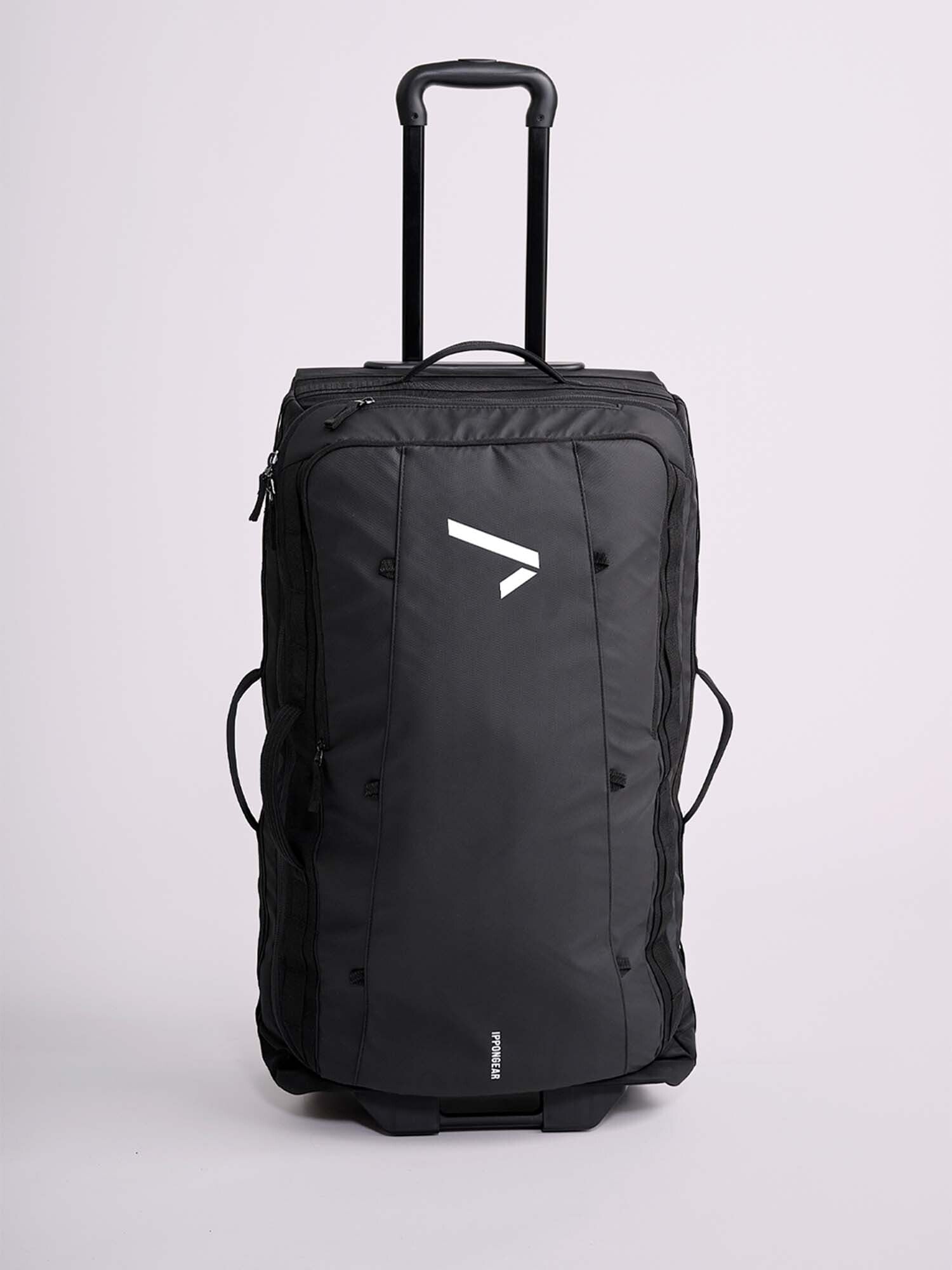 The IPPONGEAR Travel Trolley Black L is a large black wheeled duffel bag with a telescopic handle, side handles, and a white geometric logo on the front, shown upright on a plain light background.