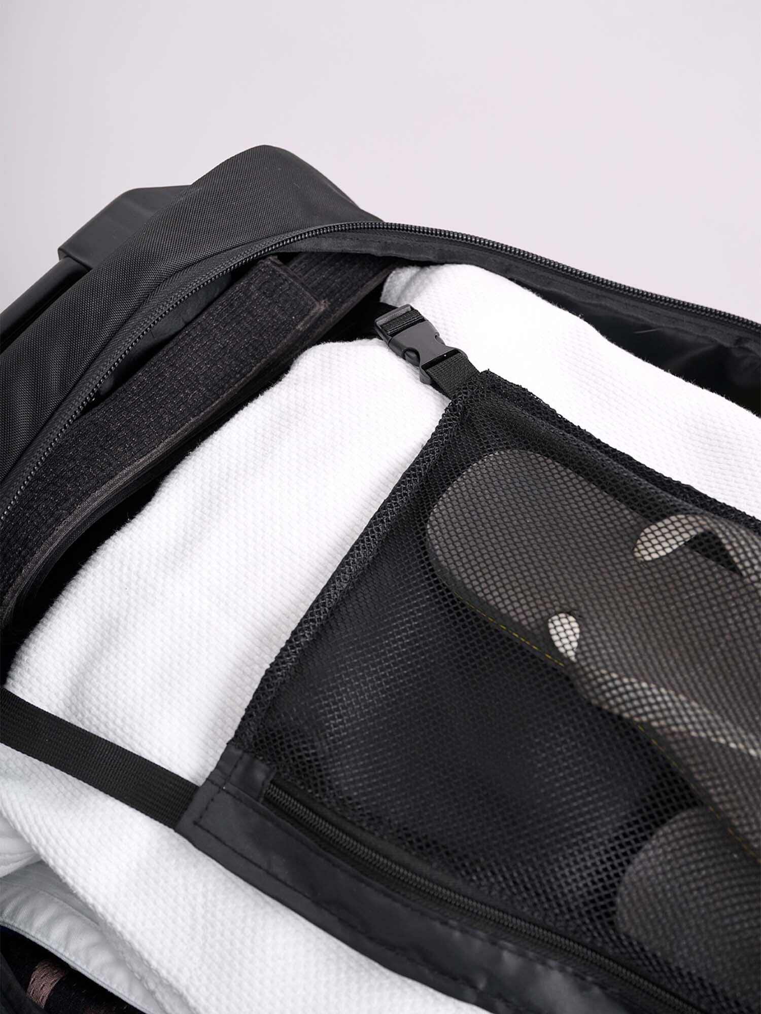 A close-up of the open IPPONGEAR Travel Trolley Black L reveals a neatly packed white towel and flip-flops inside its black mesh zippered pocket.