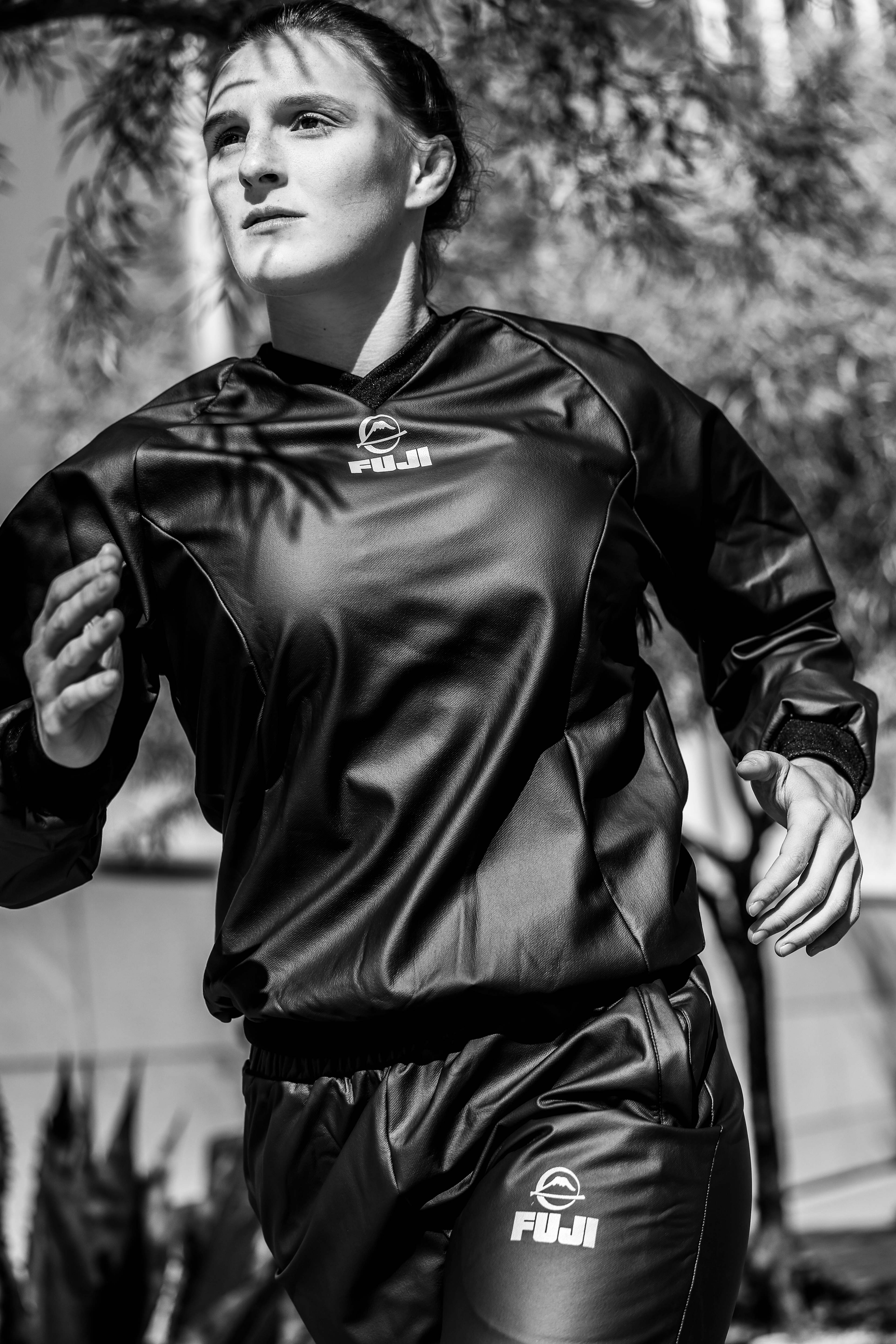 A woman in a sports tracksuit with FUJI logos runs outdoors, looking focused. Sunlight and shadows fall across her face and body, with trees and plants in the background. The image is in black and white.
