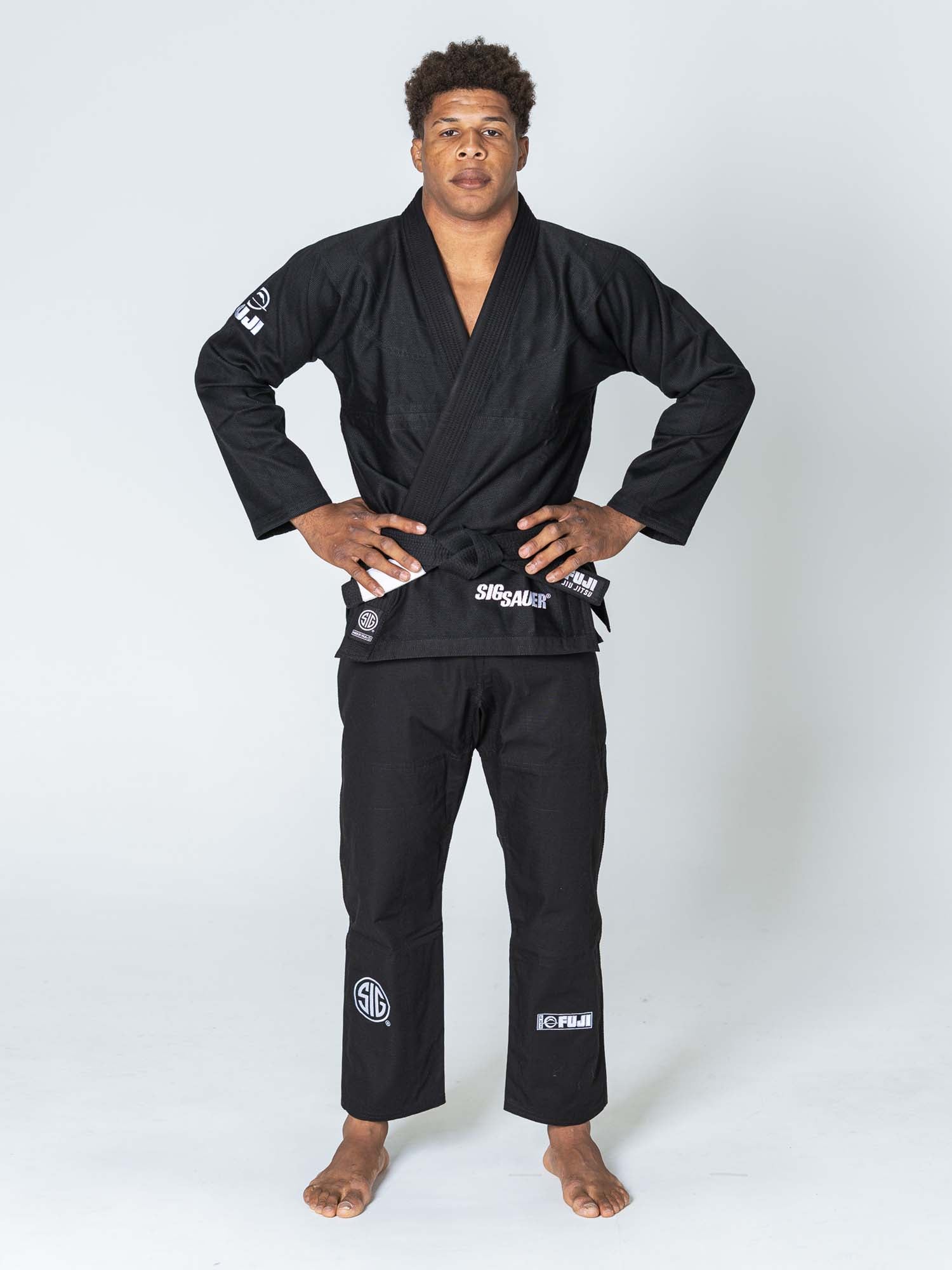 A person stands barefoot on a white background, wearing the SIG SAUER Suparaito Gi Black with white logos. Their hands are on their hips as they look straight ahead with a neutral expression.