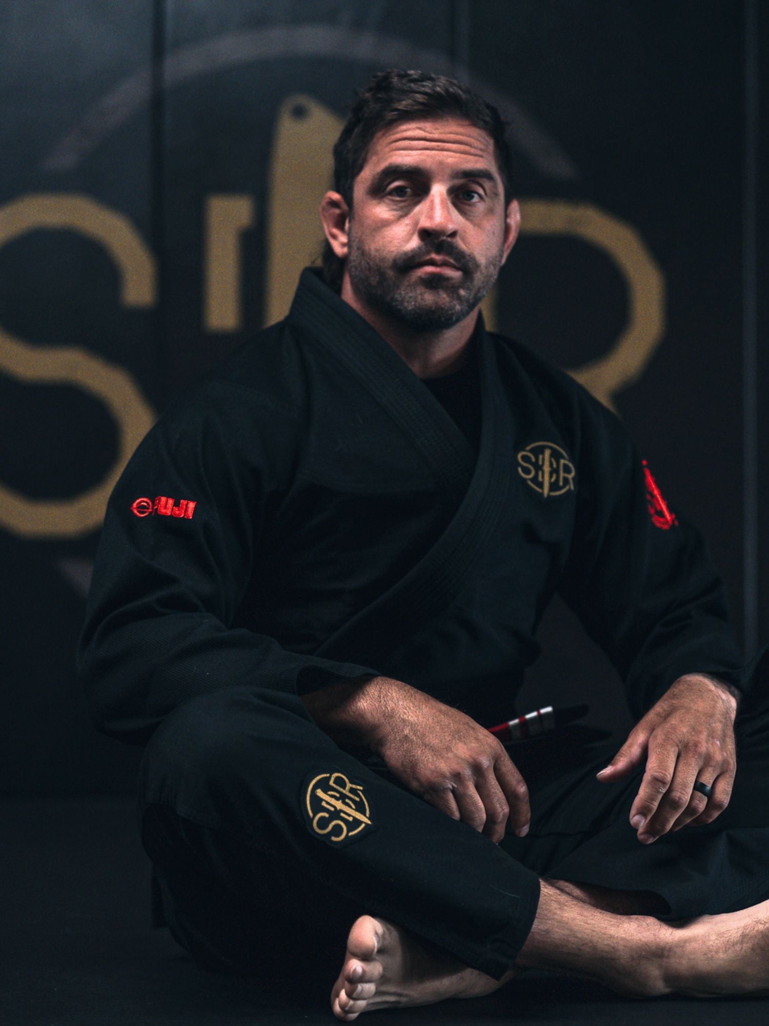 A man sits barefoot on the floor, looking thoughtful, wearing the Sheepdog Response Standard Suparaito BJJ Gi in black with gold and red patches. The background features a blurred logo.
