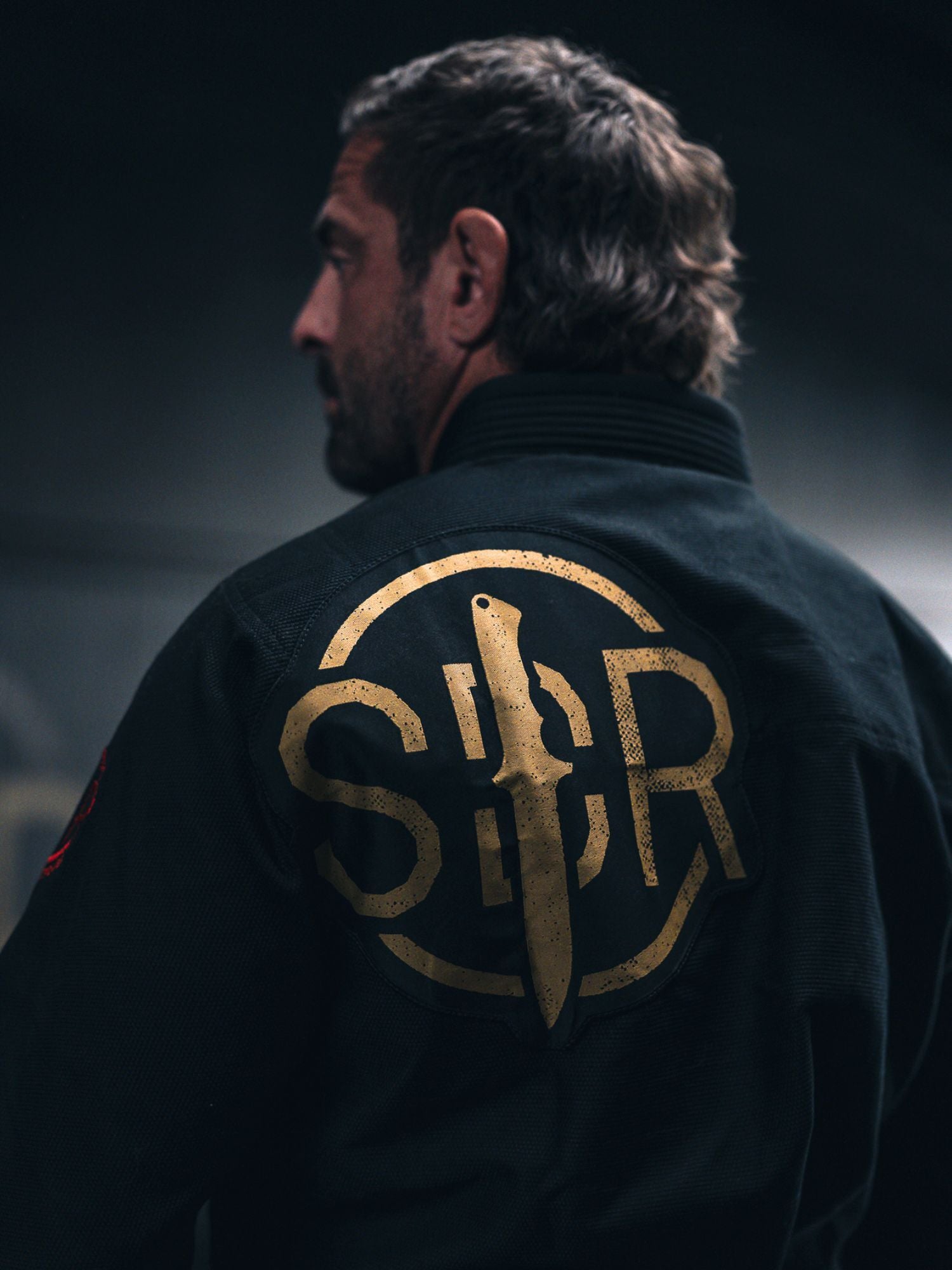 A person is seen from behind wearing the Kids Sheepdog Response Standard Suparaito BJJ Gi in black, which features a prominent gold “SBR” logo and knife design on the back. The background is dark and blurred.