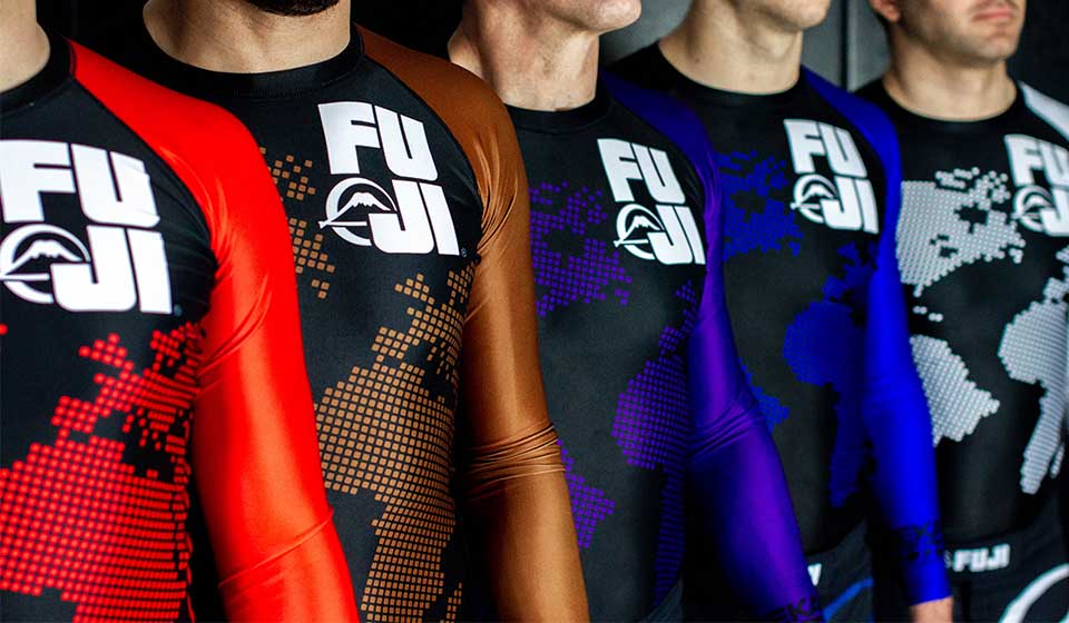 Five people wearing FUJI branded rash guards stand side by side. Each rash guard has a different color sleeve, representing various Brazilian Jiu-Jitsu belt ranks: white, blue, purple, brown, and black.