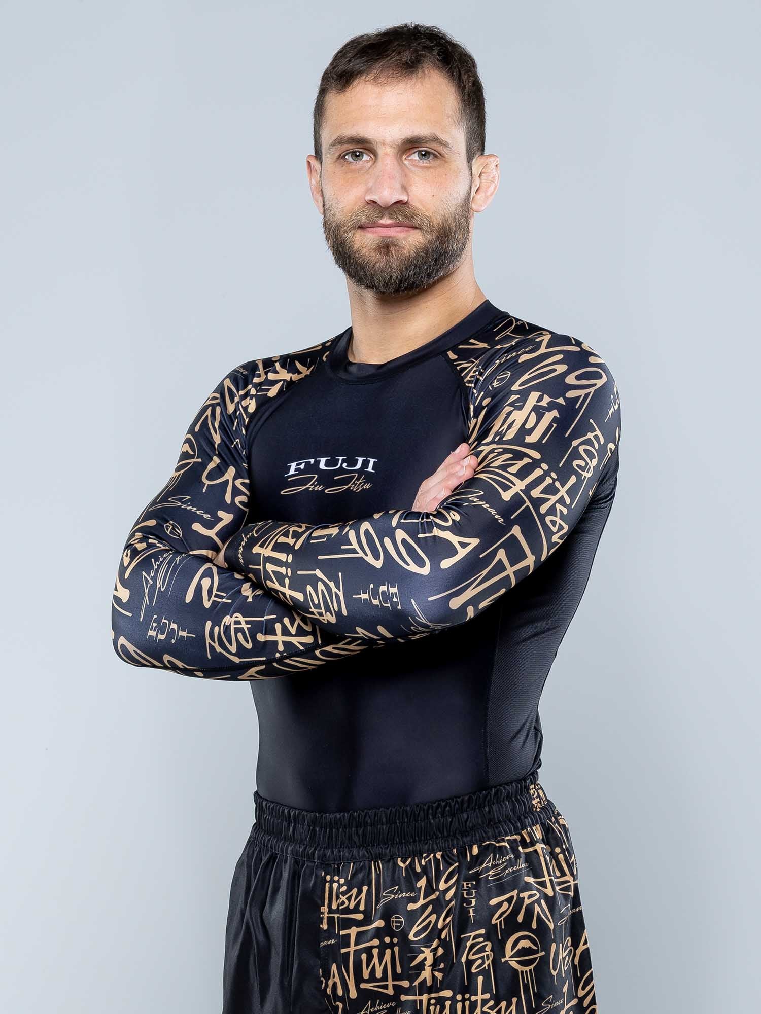 A man with short brown hair and a beard stands with arms crossed, wearing the Shinobi Long Sleeve Rashguard Black and matching shorts with gold graffiti designs, against a plain light gray background.