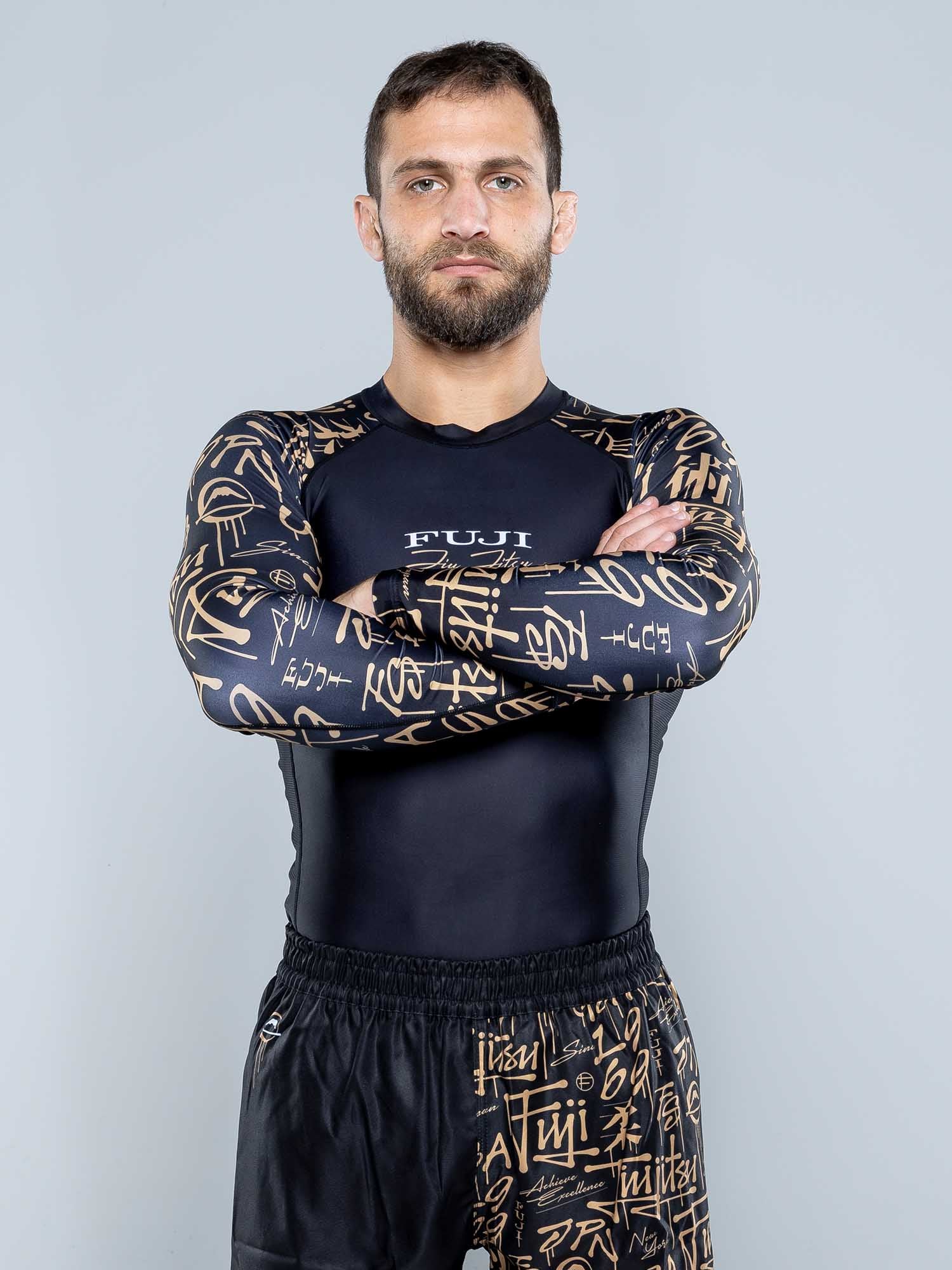A bearded man stands with arms crossed, wearing the Shinobi Long Sleeve Rashguard Black and matching shorts against a plain light gray background.