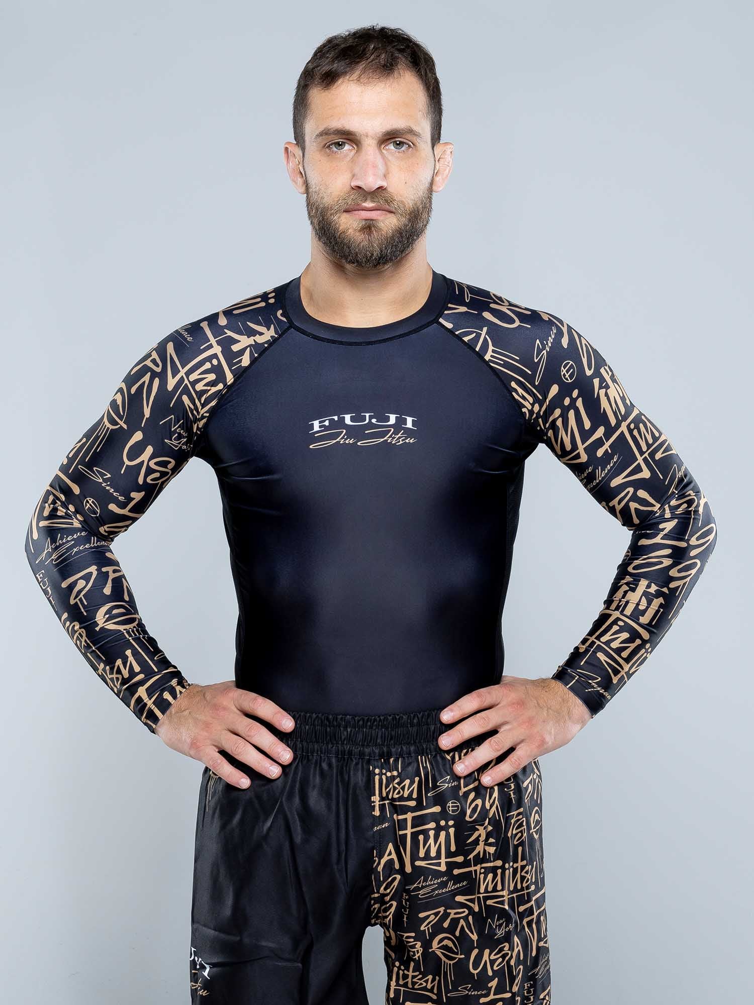 A man with short brown hair and a beard stands facing forward, hands on hips, wearing a black Shinobi Long Sleeve Rashguard and matching athletic pants with gold graffiti-style writing against a plain light gray background.