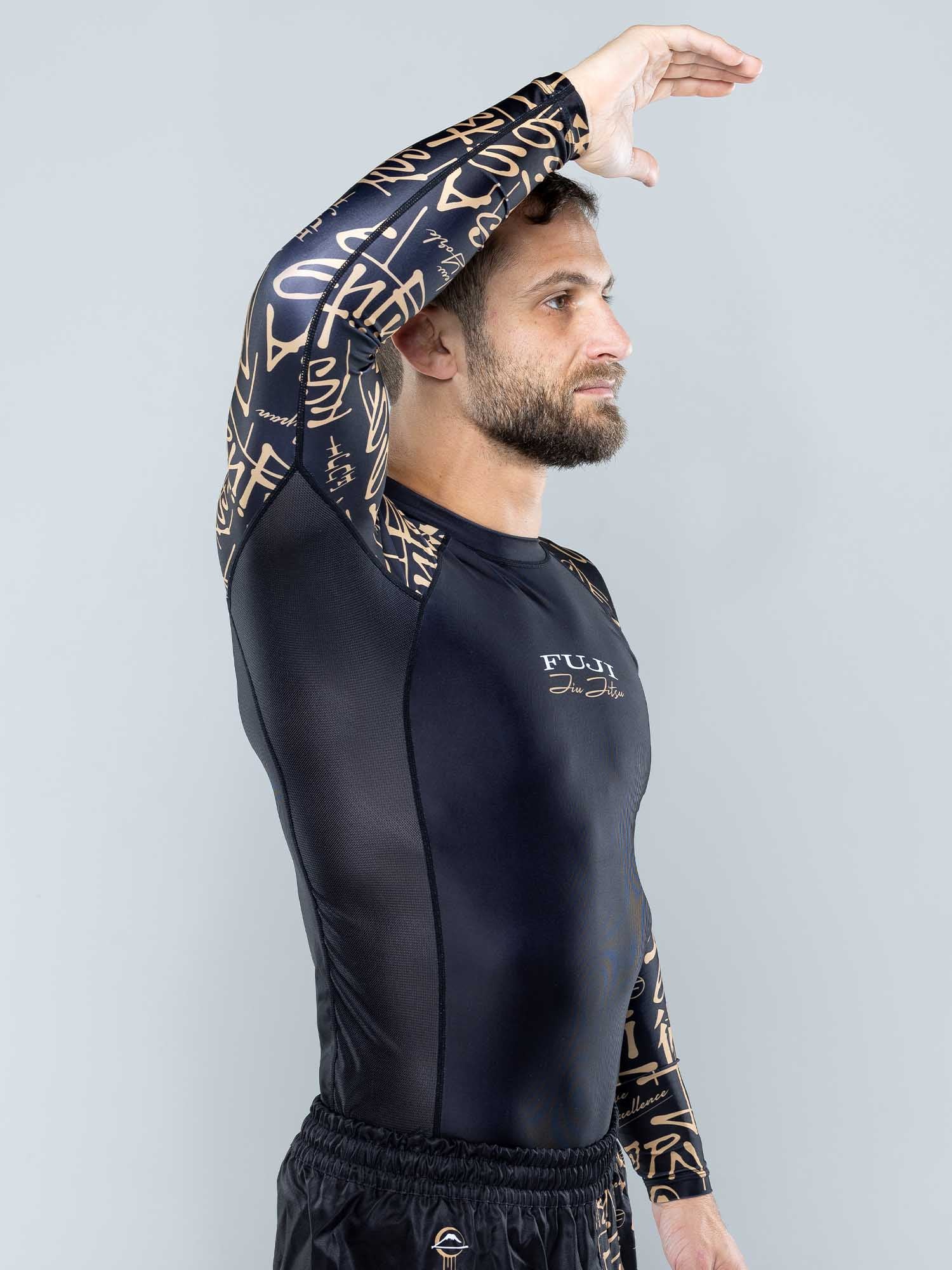 A man in the Shinobi Long Sleeve Rashguard Black stands sideways, raising his left arm over his head with a neutral expression against a plain light background.