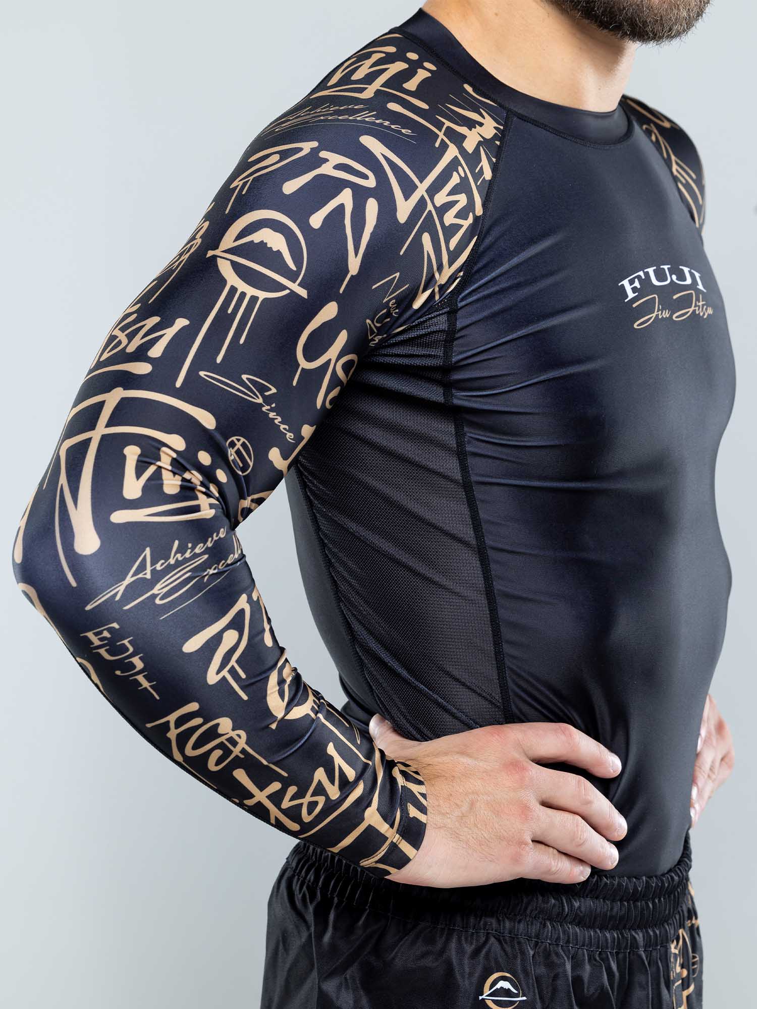 A man wearing the Shinobi Long Sleeve Rashguard Black, featuring gold graffiti-style sleeve designs and "FUJI Jiu Jitsu" on the chest, stands with his hands on hips.