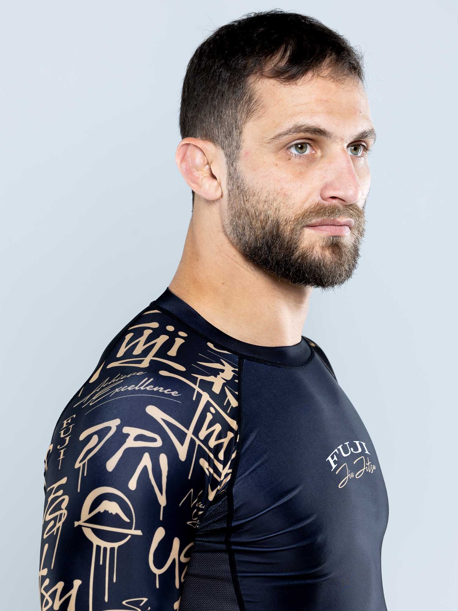 A man with short brown hair and a beard models the Shinobi Long Sleeve Rashguard Black, featuring beige graffiti-style sleeve designs, standing against a light gray background and looking to the side.