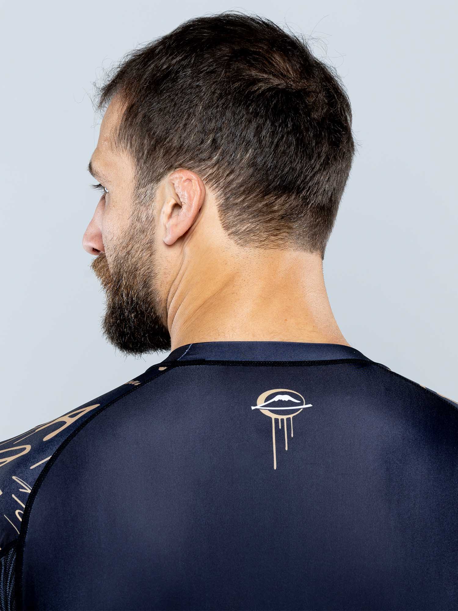 A man with short brown hair and a beard is seen from behind, wearing the Shinobi Long Sleeve Rashguard Black featuring a stylized eye with a dripping design on the upper back.