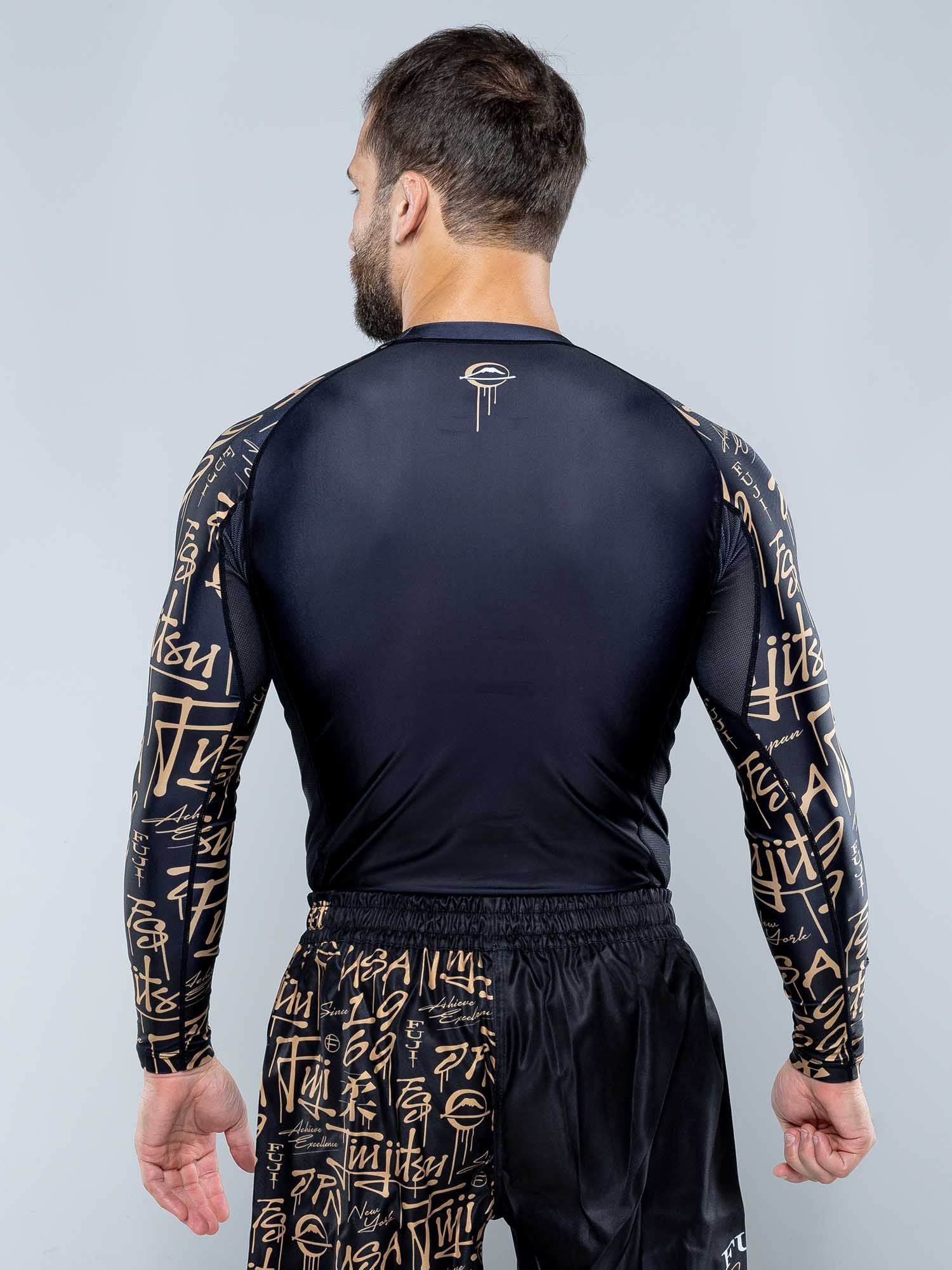 A man with short hair and a beard stands with his back to the camera, wearing the Shinobi Long Sleeve Rashguard Black and matching shorts featuring tan graffiti-style designs on the sleeves and shorts.