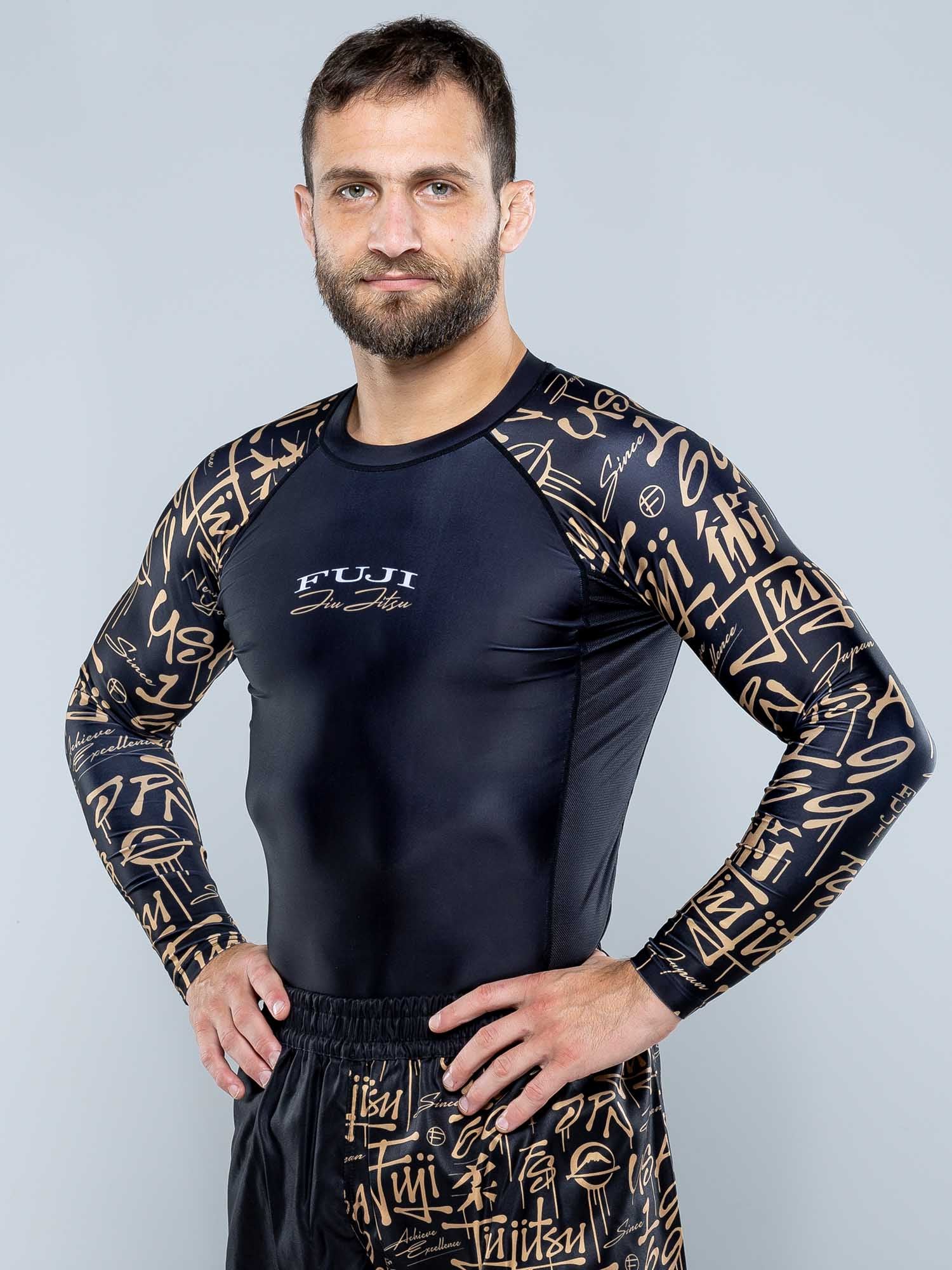 A bearded man stands confidently with his hands on his hips, wearing the Shinobi Long Sleeve Rashguard Black featuring gold graffiti-style lettering on the sleeves and matching shorts.