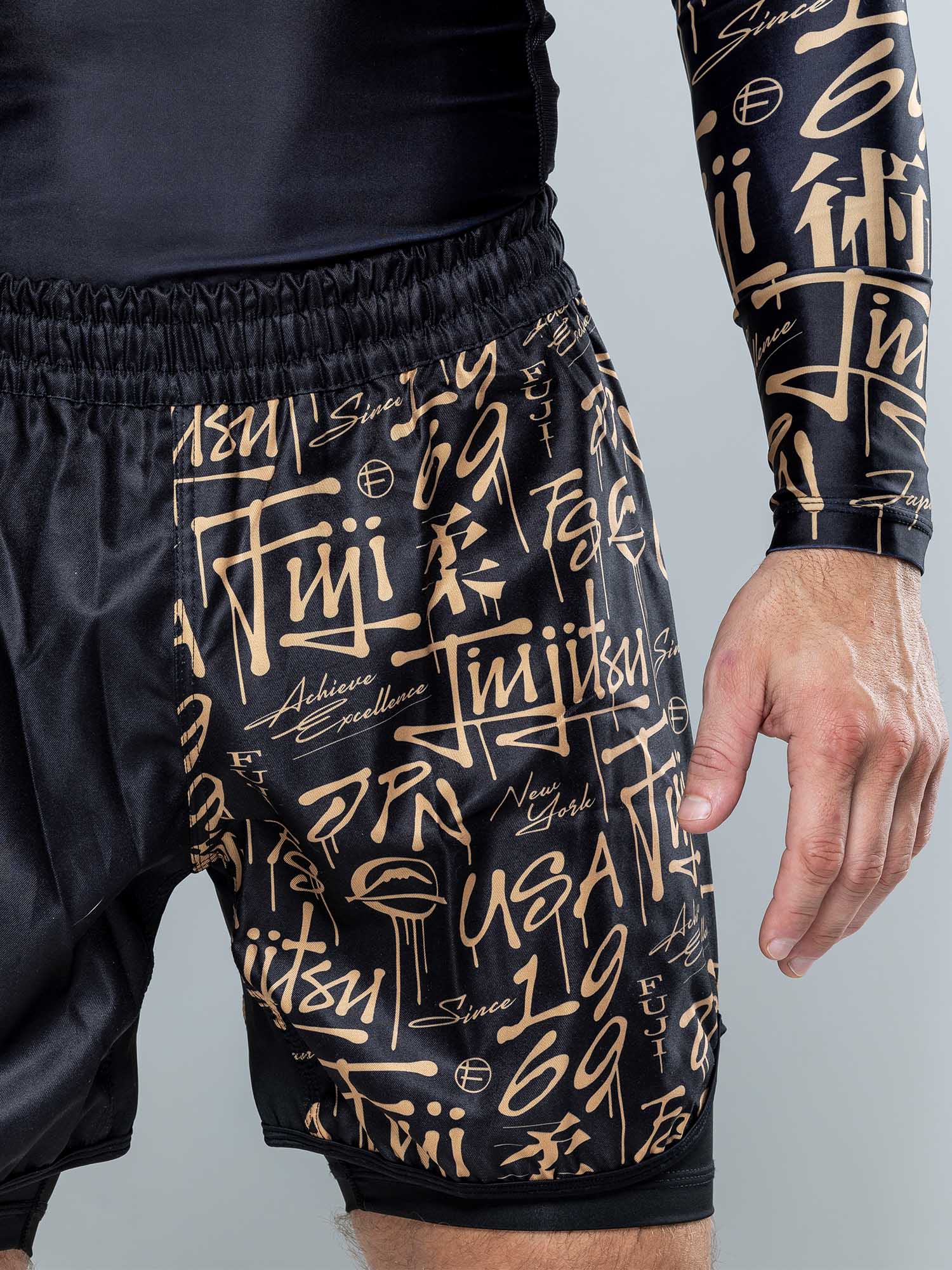 A person wearing Shinobi Fight Shorts Black With Liner and a matching long-sleeve top, both featuring gold stylized lettering, graffiti-inspired designs, the words "jiu jitsu," numbers, and abstract symbols.