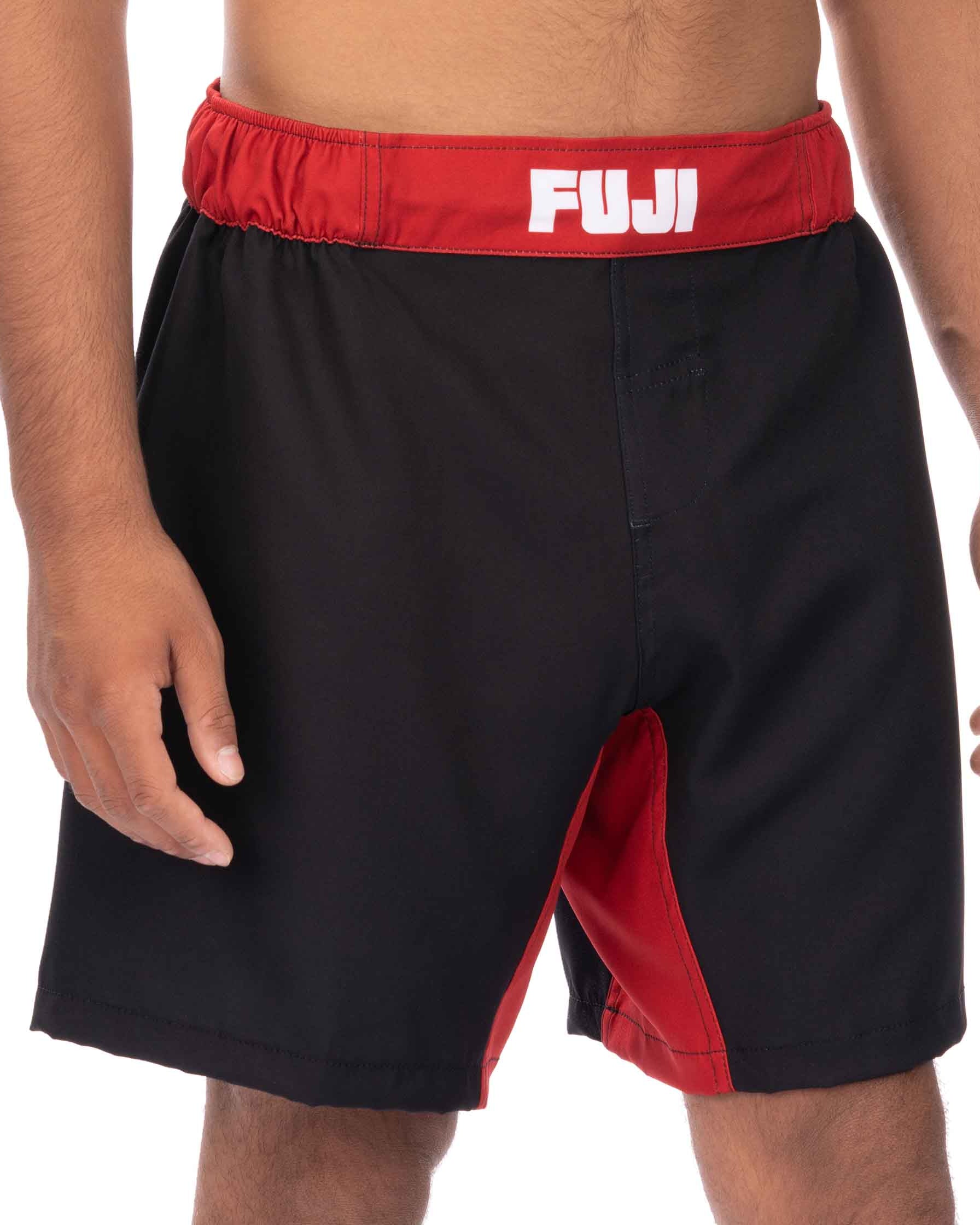 A person wears Essential Grappling Red Fight Shorts, featuring a red waistband with "FUJI" in white letters, black fabric, and red inner panels. The person's torso is partially visible.